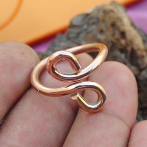 Puede incluir: Un anillo de cobre pulido con un diseño de bypass que presenta dos extremos abiertos y redondeados que se curvan hacia adentro. El metal tiene un brillo cálido y una superficie lisa y continua, formando una estructura de bucle abierto.