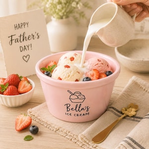 May include: A light pink ceramic bowl labeled Bella's ICE CREAM containing scoops of strawberry and vanilla ice cream with fresh strawberries, blueberries, and mint leaves. A small pitcher pours milk into the bowl, accompanied by a gold flower spoon.