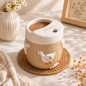 May include: A ceramic mug with a speckled tan body featuring raised white duck figures with orange beaks and feet. The top rim is white with a speckled texture and includes the name Charlotte engraved near a small circular opening for a straw.