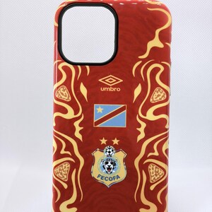Peut inclure: Une coque de smartphone rouge avec un motif tourbillonnant jaune, le logo Umbro, le drapeau de la République démocratique du Congo et l'écusson FECOFA avec une illustration de léopard et deux étoiles dorées au-dessus, centrés au dos.