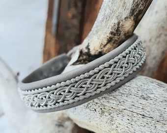 May include: A gray leather bracelet with a silver braided design. The bracelet is on a wooden surface.