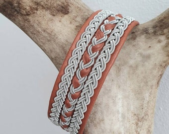 May include: A brown leather bracelet with a silver braided design. The bracelet is woven with three strands of silver cord.