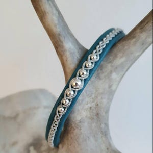 Può includere: Bracciale in pelle color teal con dettagli in metallo argentato. Il bracciale presenta una fila di perline argentate e un design argentato intrecciato. Il bracciale è esposto su un corno di cervo marrone chiaro.