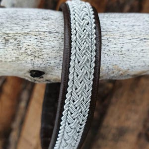 Pode incluir: Um bracelete de couro castanho com uma banda trançada prateada. O bracelete está enrolado em torno de um pedaço de madeira à deriva.