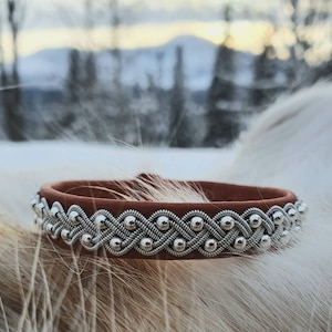Könnte beinhalten: Ein braunes Lederarmband mit einem geflochtenen Silberdesign und silbernen Perlen.