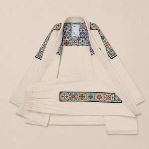 May include: An off-white martial arts gi featuring a colorful, multi-patterned tile design along the shoulders and the lower hem. The interior neck label displays the text ML. The fabric has visible stitching throughout the jacket and pants.