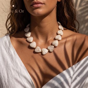 Peut inclure: Un collier composé d'une série de perles lisses en forme de cœur avec un motif marbré crème et beige, séparées par de petites perles intercalaires dorées. Le texte "Nacre & Or" est visible dans le coin supérieur gauche de l'image.