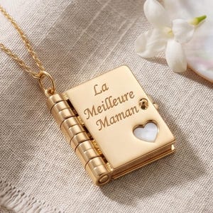 Peut inclure: Un pendentif en métal doré en forme de livre miniature, suspendu à une chaîne assortie. La couverture porte l'inscription gravée 'La Meilleure Maman' et une petite découpe en forme de cœur révélant une incrustation blanche irisée.