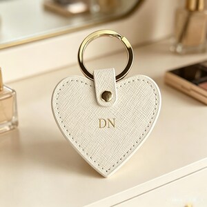 May include: A heart-shaped keychain made of off-white textured material with gold-tone hardware. The center features the gold-embossed letters DN. A gold metal ring is attached to the top by a small tab secured with a gold-tone metal rivet.