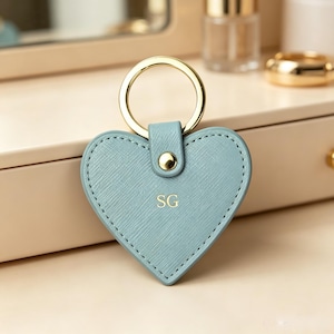May include: A heart-shaped keychain in a light blue textured material with gold-tone stitching around the perimeter. The keychain features a gold-tone metal ring attached by a small tab with a gold-tone rivet. The letters SG are embossed in gold.
