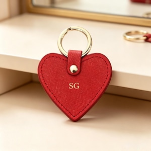 May include: A heart-shaped red keychain with a textured surface, featuring a gold-toned metal ring and a central gold-toned rivet. The letters SG are embossed in gold in the center of the heart, which is attached to the ring by a small leather tab.