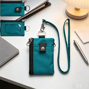 May include: A teal nylon lanyard wallet featuring a zipped main compartment, a front pocket with a snap-button closure, and a detachable neck strap. The fabric has a subtle grid pattern, and the wallet includes a clear ID window on the back.