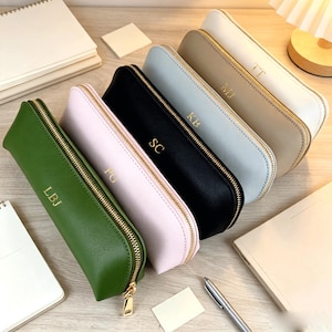 Pode incluir: Uma coleção de seis estojos retangulares nas cores verde-oliva, rosa claro, preto, azul claro, bege e branco. Cada estojo possui um zíper dourado e iniciais gravadas em dourado na parte frontal, incluindo LBJ, PU, SC, KB, MJ e TT.