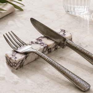 Puede incluir: Un reposacuchillos rectangular de mármol blanco con vetas de color púrpura oscuro, que presenta un tenedor de metal plateado y un cuchillo a juego con un mango texturizado y martillado, apoyados horizontalmente sobre el bloque.