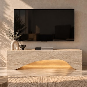 Op de afbeelding: Een rechthoekig beige stenen tv-meubel met een gladde, gebogen uitsparing in de basis en geïntegreerde warme verlichting aan de onderzijde. Het meubel heeft een natuurlijke, gevlekte textuur en staat onder een grote flatscreen-televisie.