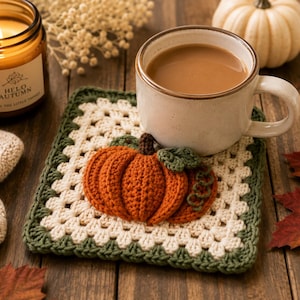 May include: A square crochet coaster featuring a cream-colored granny square pattern with a dark green border. A three-dimensional orange pumpkin with a brown stem and green leaves is attached to the center of the coaster, supporting a ceramic mug.