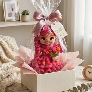 May include: A crocheted doll with pink yarn hair in a long braid, wearing a pink dress decorated with small orange and yellow flowers. The doll holds a pink crocheted flower and is packaged in a clear plastic bag with a pink ribbon and a gift tag.