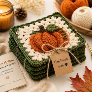 May include: A stack of square crochet coasters featuring a central orange pumpkin motif with a green stem and vine, bordered by cream and dark green yarn. A brown paper tag tied with twine reads "Handmade with heart" alongside a crochet pattern booklet.