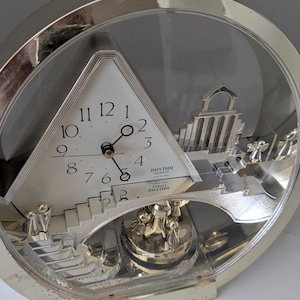 May include: A round Rhythm Quartz clock with a silver-tone frame and a triangular white face featuring black numerals. The clock includes a decorative silver-tone architectural scene with stairs and small figures, all encased behind a clear circular cover.