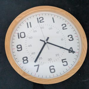 Op de afbeelding: Een ronde wandklok met een licht houten frame en een witte wijzerplaat. De klok heeft grote zwarte Arabische cijfers van 1 tot 12, met een secundaire binnenring van kleinere getallen van 05 tot 60. De klok heeft drie zwarte wijzers.