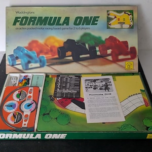Könnte beinhalten: Waddingtons Formula One Autorennen-Brettspiel. Der Deckel zeigt bunte Plastik-Rennwagen. Die geöffnete Schachtel enthält ein Spielbrett mit Streckenillustration, eine Strafentabelle, eine Spielanleitung, Würfel und eine Armaturenbrett-Konsole.