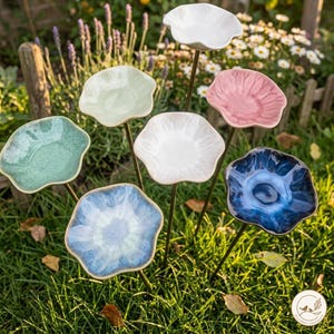 May include: A collection of seven ceramic flower-shaped garden stakes, each featuring a unique glazed finish in colors including white, light green, pink, dark blue, and light blue, mounted on thin metal rods for placement in the ground.