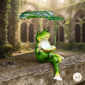 May include: A green frog figurine sitting and reading a small book while holding a large, dark green leaf overhead as an umbrella. The frog has a white belly, yellow-rimmed eyes, and a smooth, glossy finish with visible water droplets.