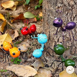 May include: A set of five metal ant sculptures in bright colors including red, orange, blue, green, and purple. Each ant features a round head with two eyes, a segmented body, and thin wire legs. The purple ant is positioned on a tree trunk.