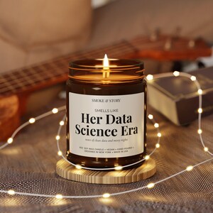 Könnte beinhalten: Eine brennende Sojawachskerze in einem bernsteinfarbenen Glas mit einem weißen Etikett, das SMOKE & STORY, SMELLS LIKE Her Data Science Era, notes of clean data and messy nights, 8oz, vegan, hand-poured, designed in New York, made in USA, zeigt.