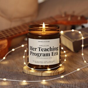Op de afbeelding: Een kaars in een bruine glazen pot met een wit etiket met de tekst Smoke & Story, Smells Like Her Teaching Program Era, notes of a first classroom, waiting, 9oz soy wax candle, vegan, hand-poured, designed in New York, made in USA.