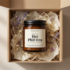 May include: A 255g soy wax candle in an amber glass jar with a black lid, nestled in a cardboard gift box with crinkle paper and dried lavender. The label reads: SMOKE & STORY, SMELLS LIKE Her PhD Era, notes of a dissertation and stubbornness.