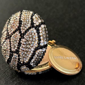 May include: A round Estée Lauder compact case featuring a basketball design covered in clear and black crystals. The gold-tone metal compact is partially open, revealing a gold interior surface with the Estée Lauder brand name printed on it.