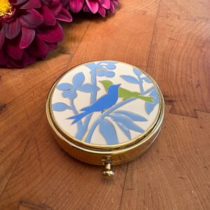 May include: A round gold-tone metal compact case featuring an enamel inlay on the lid. The design displays a white background with light blue and lime green stylized birds perched on branches with light blue leaves and berries.