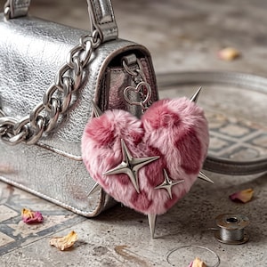 May include: A metallic silver handbag with a thick chain handle features a heart-shaped pink faux fur bag charm. The plush heart is decorated with silver-toned metal star-shaped accents and sharp spikes, attached to the bag by a heart-shaped clasp.