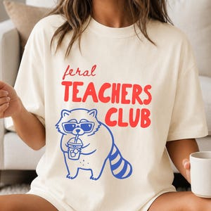 Puede incluir: Una camiseta de color crema con el texto rojo "feral TEACHERS CLUB" sobre un gráfico azul de un mapache que lleva gafas de sol y sostiene una bebida helada. El diseño está centrado en la parte delantera de la camiseta de manga corta.
