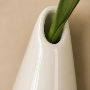 以下が含まれることがあります： 滑らかな白い陶器製の花瓶で、先細りのしずくのようなシルエットをしており、上部に楕円形の開口部が一つあります。鮮やかな緑色の植物の茎と、2枚の長く尖った葉が、光沢のある白い容器の開口部から上に向かって伸びています。