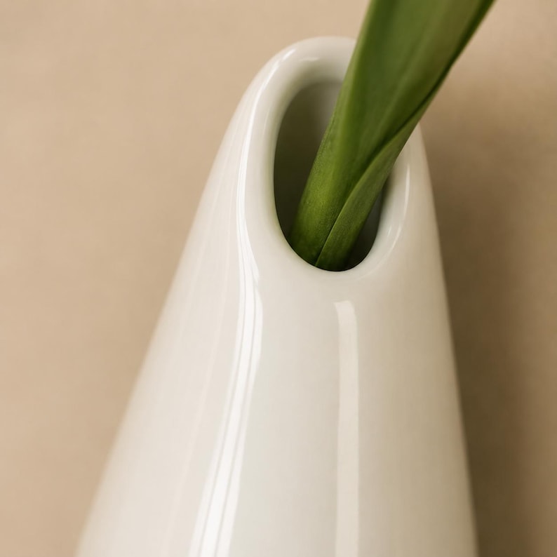 以下が含まれることがあります： 滑らかな白い陶器製の花瓶で、先細りのしずくのようなシルエットをしており、上部に楕円形の開口部が一つあります。鮮やかな緑色の植物の茎と、2枚の長く尖った葉が、光沢のある白い容器の開口部から上に向かって伸びています。