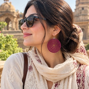 May include: A pair of circular, deep magenta earrings featuring a detailed, laser-cut geometric pattern of concentric circles and small perforations. The earrings hang from silver-toned hooks and are worn against a light-coloured, patterned fabric.