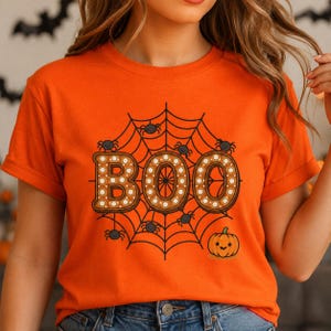 Puede incluir: Una camiseta naranja de manga corta con un estampado gráfico de una telaraña negra y la palabra BOO en letras grandes de color marrón con puntos blancos. Varias arañas pequeñas negras están sobre la red y una calabaza está debajo.