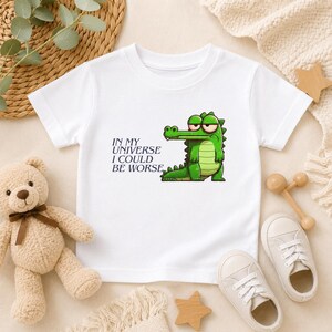 May include: A white short-sleeved t-shirt featuring a graphic of a green cartoon crocodile with a grumpy expression. Next to the crocodile, the text reads, IN MY UNIVERSE I COULD BE WORSE, printed in a dark blue, serif font on the front of the shirt.