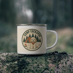 May include: A white enamel mug featuring a circular graphic with a tan dog wearing a bandana, a tent, a campfire, and pine trees. The text "CAMP COCKAPOO" is printed in a dark green arched font along the top of the circular design.