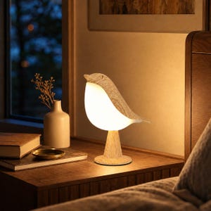 Puede incluir: Una lámpara de mesa con forma de pájaro que presenta una textura de veta de madera clara en la cabeza y la cola, con un vientre blanco liso y brillante que emite una luz cálida. La lámpara se apoya sobre una base circular de madera.