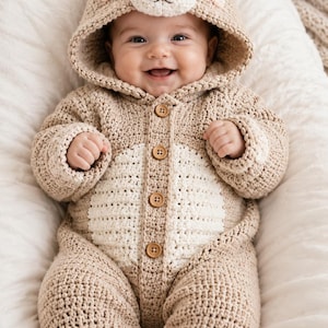 May include: A beige crochet baby onesie featuring a hood with bear ears, a white belly panel, and four brown wooden buttons down the front. The feet of the onesie have white paw pad details, and the hood includes a bear face with black eyes.