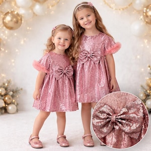 May include: Two dresses in shimmering rose gold sequins, featuring short sleeves with ruffled edges, a round neckline, and a large decorative bow centred at the waist. The fabric is fully covered in small, reflective sequins that catch the light.