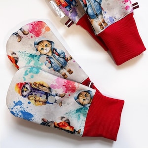 May include: A pair of mittens featuring a light gray background with a colorful watercolor splatter pattern. The print depicts cartoon animals wearing winter clothing, including cats in hoodies and an owl in a hat. The mittens have bright red ribbed cuffs.