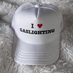 Può includere: Un cappellino trucker bianco con retro in rete e pannello frontale rigido. La parte anteriore presenta il testo "I" seguito da un simbolo di cuore rosso e la parola "GASLIGHTING" stampata in grassetto con lettere maiuscole nere.