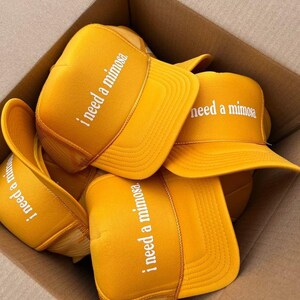May include: A collection of bright yellow trucker caps featuring a foam front panel and mesh back. Each cap is printed with the white text "i need a mimosa" in a lowercase sans-serif font. The caps are stacked together in a cardboard box.