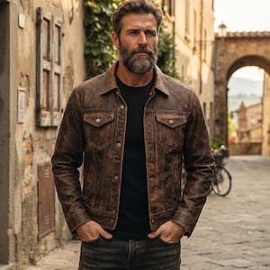 Può includere: Una giacca in pelle marrone dall'aspetto vissuto con colletto a punta, maniche lunghe e due tasche sul petto con patta e bottone. La giacca presenta una chiusura frontale con bottoni ed è indossata sopra una t-shirt nera con jeans grigio scuro.
