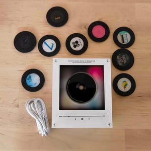 May include: A white square music player display with a central black disc, accompanied by nine circular discs featuring various graphic designs and text, and a white power cable. The display features a digital interface with playback controls.