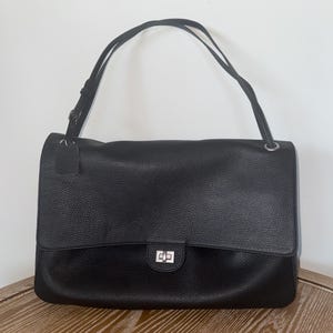Puede incluir: Un bolso de hombro de cuero negro con textura granulada, que presenta una solapa rectangular, un cierre giratorio rectangular de tono plateado y una correa de hombro fina y ajustable sujeta con herrajes plateados y una pequeña etiqueta de cuero.